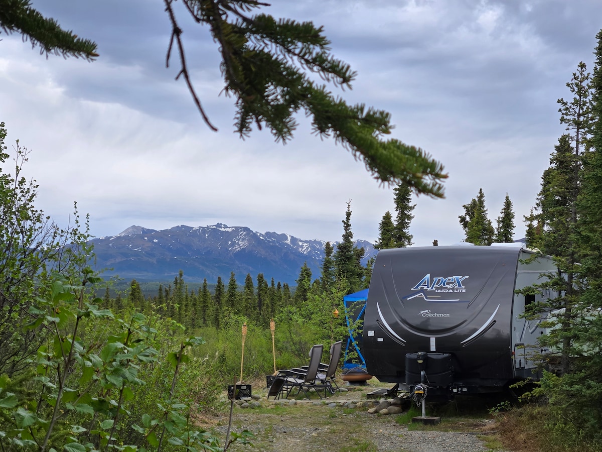 A camper trailer is positioned on a gravel area surrounded by lush green foliage. The backdrop features distant mountains, partially covered by clouds. Two outdoor chairs are visible beside a blue frame, enhancing the serene, secluded environment.