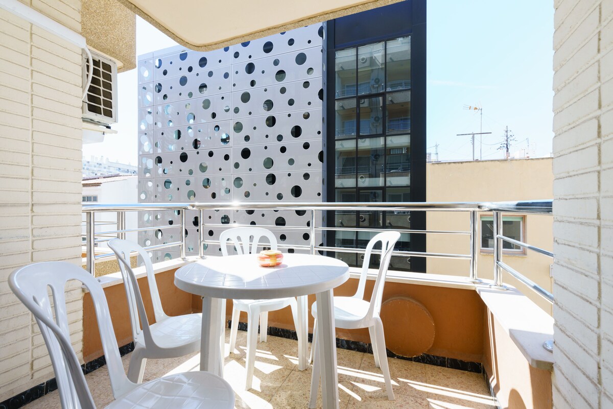 An outdoor balcony is furnished with a round table and four white chairs. A view is provided of a neighboring building with a distinctive perforated façade. Bright daylight enhances the space, creating an open and airy atmosphere.