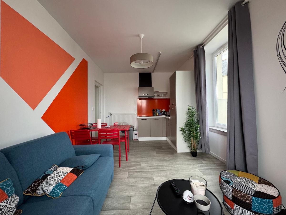 A bright living area features a spacious blue couch with colorful decorative pillows. A modern red dining table with four chairs is positioned nearby. The open kitchen is visible in the background, showcasing contemporary appliances. A small indoor plant adds a touch of greenery.