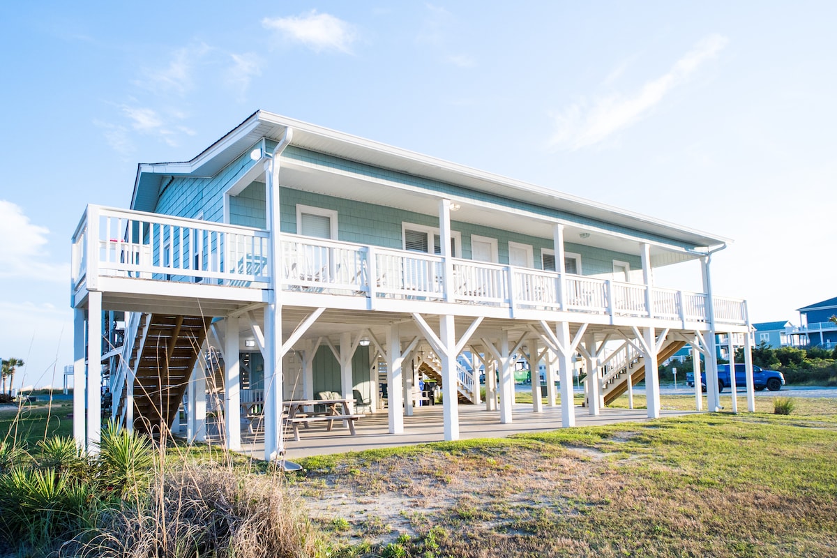 The exterior of a two-story house is displayed, featuring a light blue façade and a spacious deck with white railings. Steps lead to the entrance, and seating areas are seen on the deck. The surrounding yard is grassy, enhancing the property’s appeal.