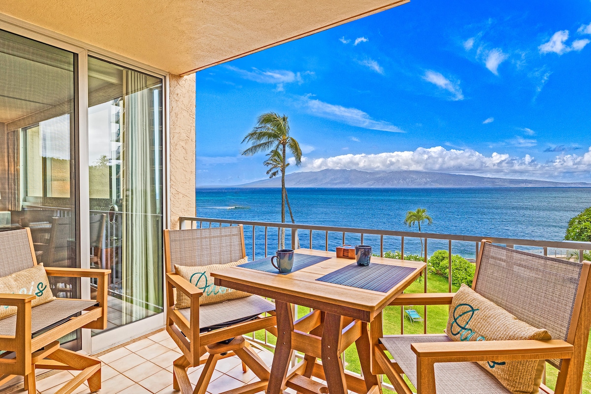 A private lanai features a wooden table and two chairs, overlooking the serene ocean and distant island views. Palm trees sway gently in the breeze, while large glass doors allow for natural light to fill the space. The horizon showcases a vibrant blue sky.
