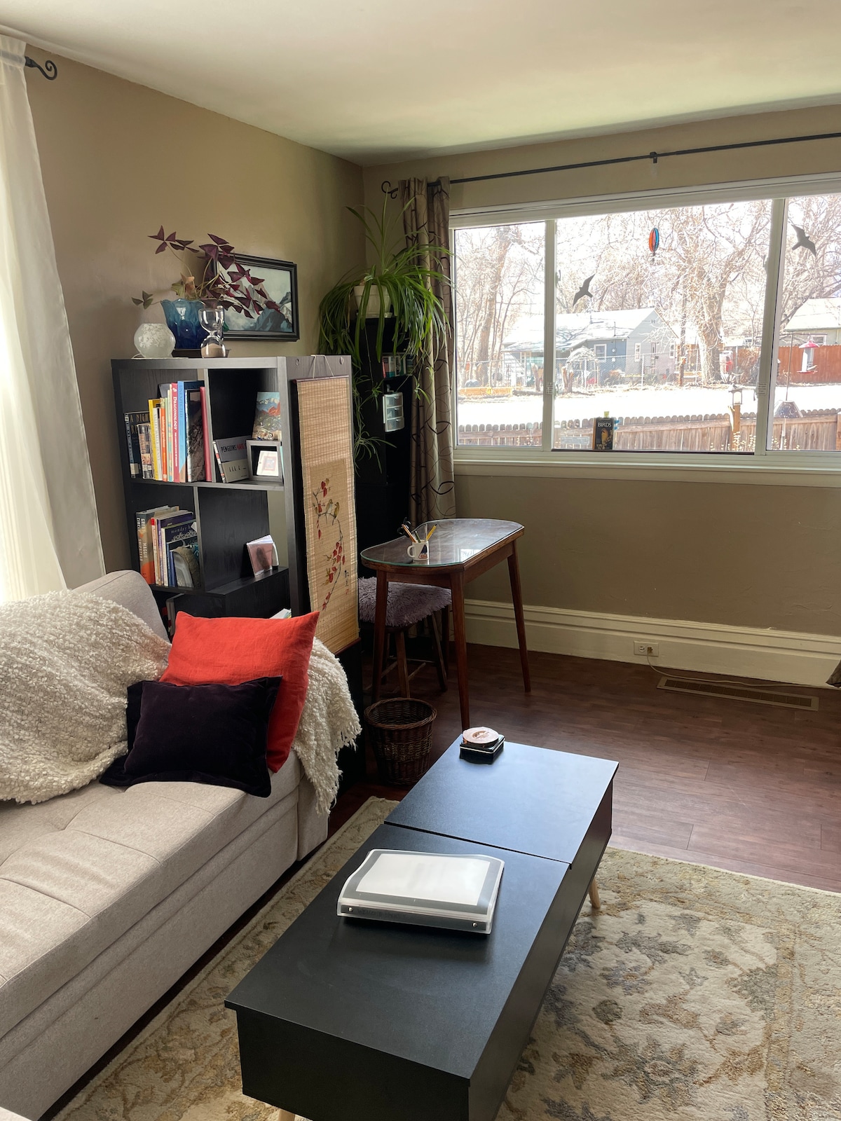 A cozy living room features a light gray sofa adorned with red and dark purple pillows. A black coffee table is placed in front, with bookshelves on one side displaying various books and decorative elements. Large panel windows allow natural light to fill the space, with views of the outdoor surroundings.