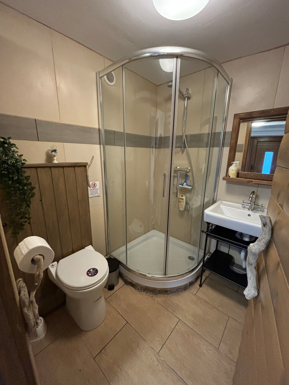 The bathroom features a glass shower enclosure, a white sink with a modern faucet, and a standard toilet. A wooden partition on one side adds warmth to the space. A mirror above the sink reflects the light, while a small plant provides a touch of greenery.