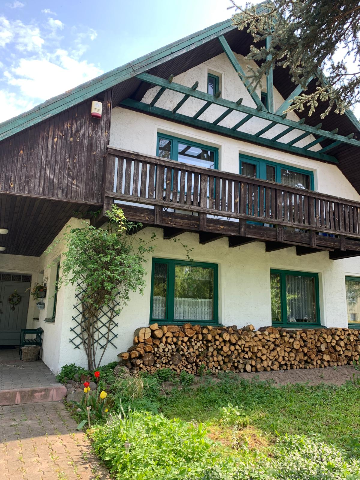 A charming two-story house features a wooden balcony supported by green beams. A stack of firewood is neatly arranged at the base, complemented by greenery. The exterior is painted white, with vibrant green window frames enhancing its traditional appearance.