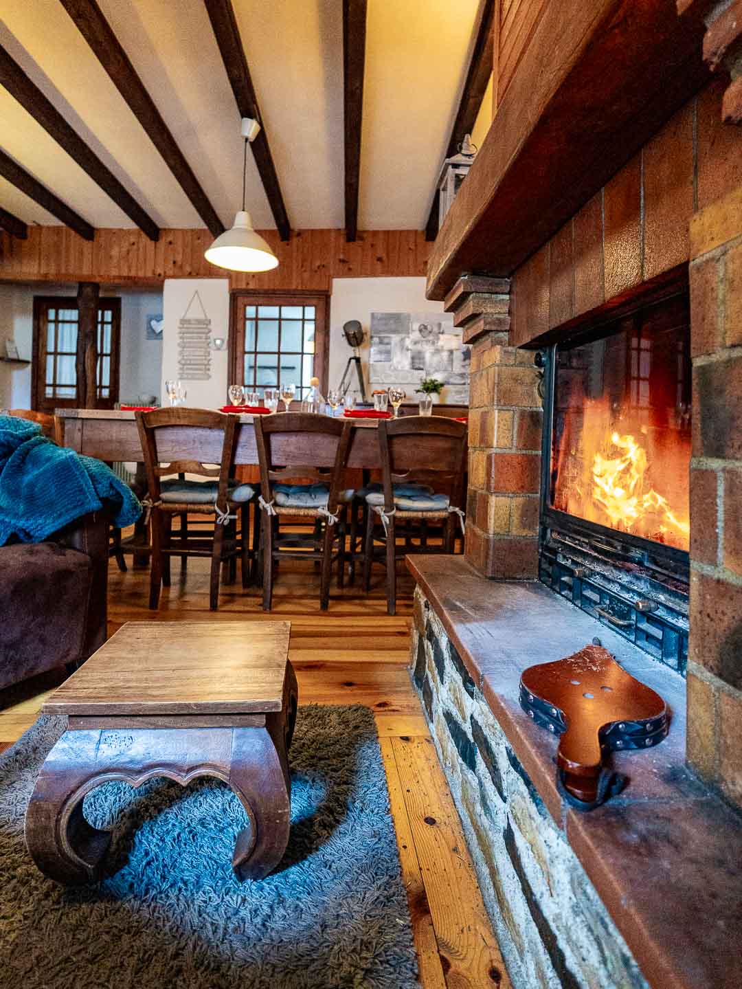 A warm living area features an inviting fireplace made of stone, with flames visible. Wooden beams decorate the ceiling above a dining table surrounded by several chairs. A small, intricately carved coffee table sits on a plush rug in the foreground.