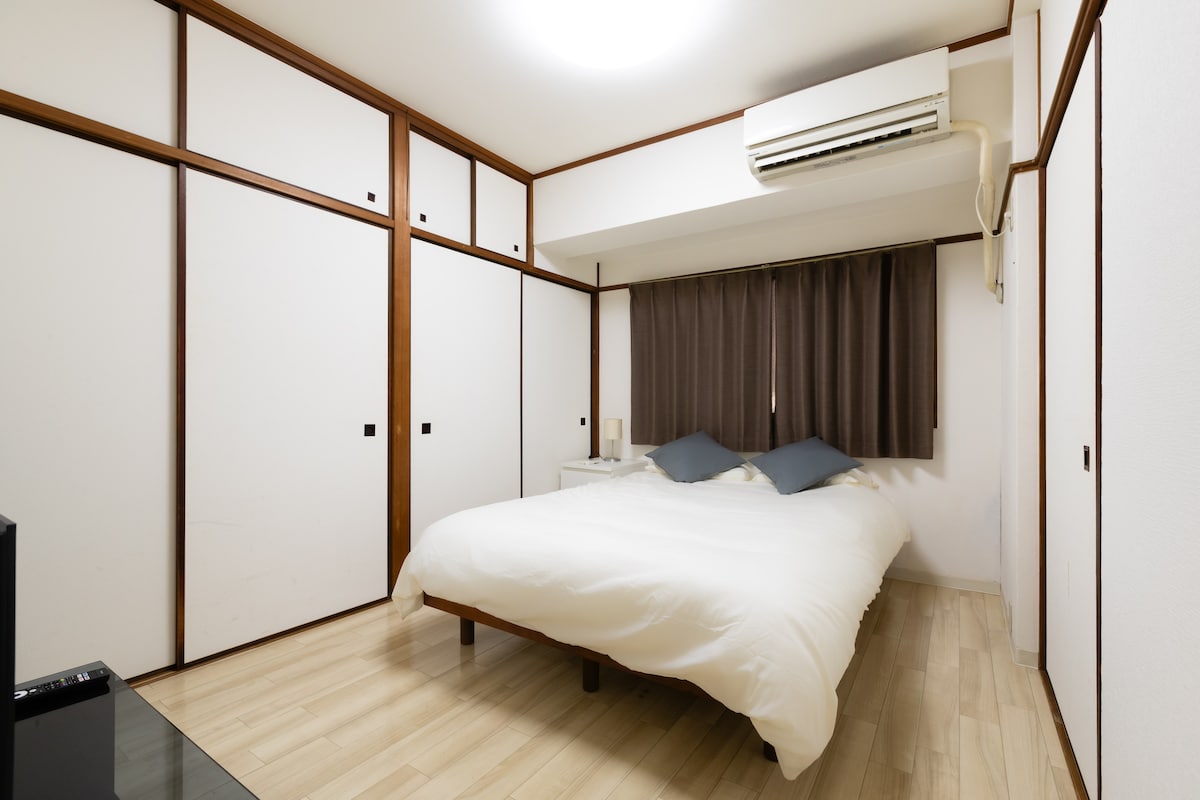 A spacious sleeping area features a soft, inviting bed adorned with neutral-toned bedding. The room is framed by sliding closet doors and is illuminated by a bright ceiling light, while dark curtains provide privacy from the outside.