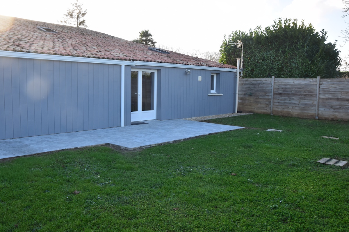 A modern, single-story building is shown with a grey exterior and a tiled roof. A concrete patio extends from the structure onto a green lawn. Fenced areas are visible on either side, providing privacy and a sense of openness.