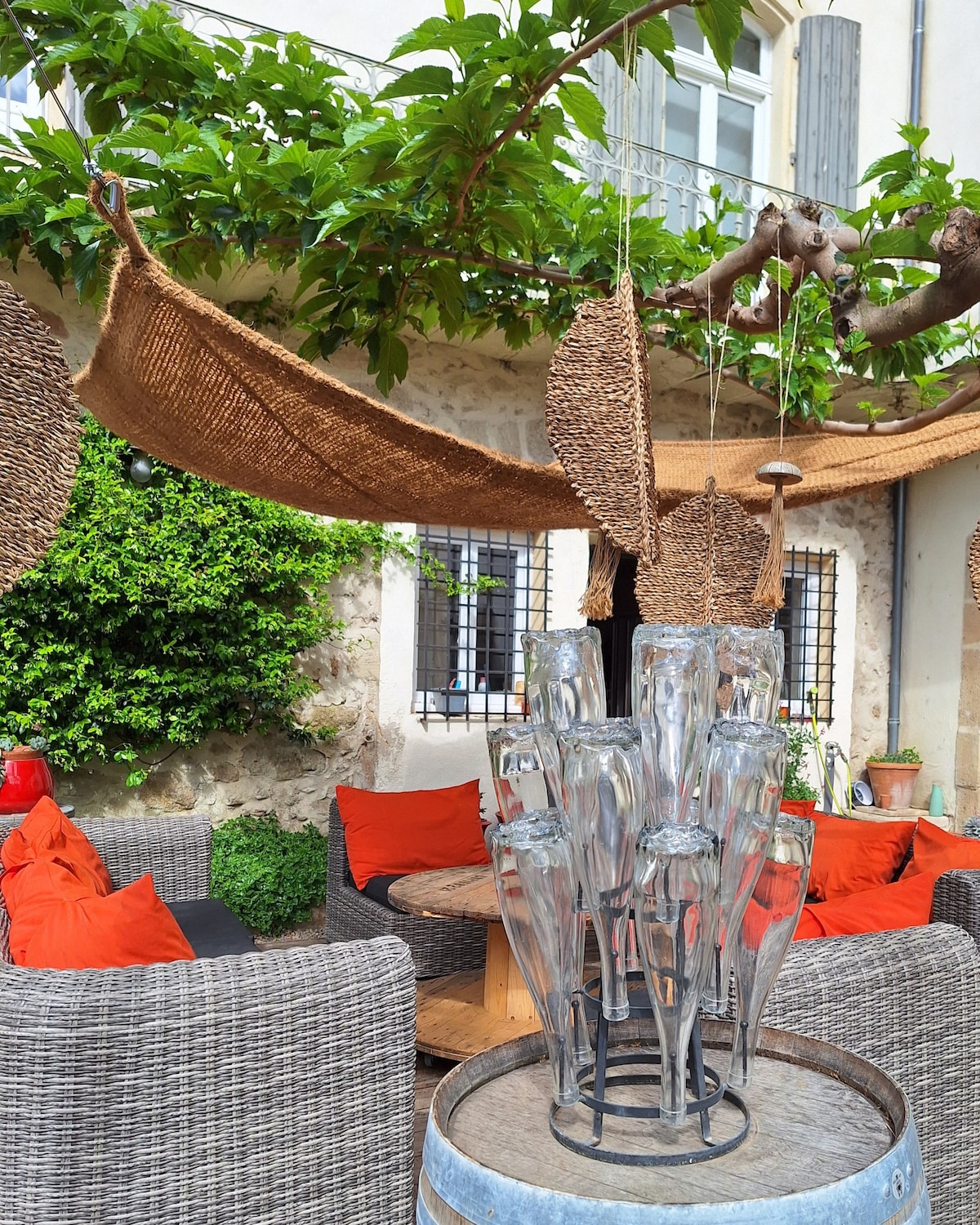 A shared outdoor seating area features woven furniture adorned with vibrant orange cushions. Strings of glassware are displayed atop a wooden barrel, and greenery is visible throughout, creating a relaxed atmosphere. A charming wall is partially seen in the background, enhancing the space's character.