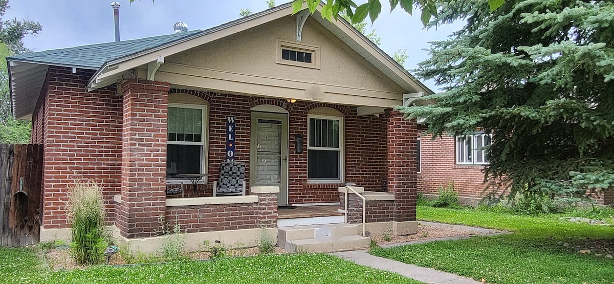 A charming brick home features a covered front porch with seating and decorative accents. Lush green grass surrounds the entrance, complemented by shrubs and trees. The entrance showcases steps leading to the porch, while the windows provide a glimpse into the welcoming interior.