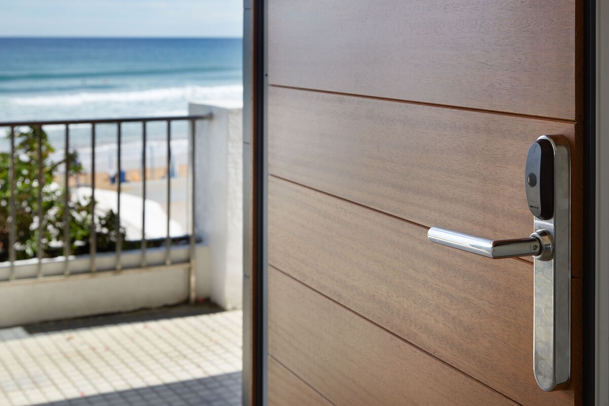 The image captures the entrance door of the apartment, showcasing a sleek wooden surface and a modern door handle. Through the open door, a view of the sea and sandy beach is visible, framed by a railing and a glimpse of outdoor space.