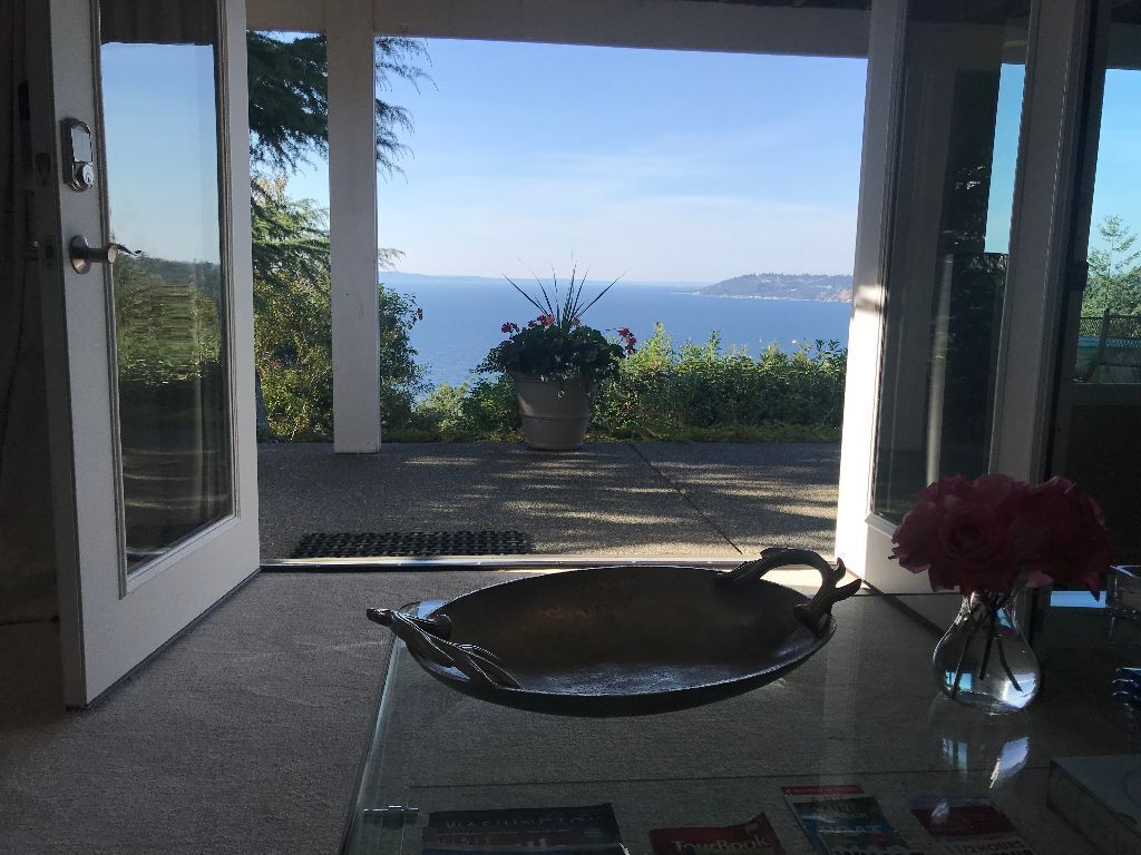 A view of Puget Sound and the surrounding greenery is captured through wide glass doors. In the foreground, a decorative metal tray rests on a glass coffee table, alongside a glass vase with pink flowers, contributing to a serene ambiance.