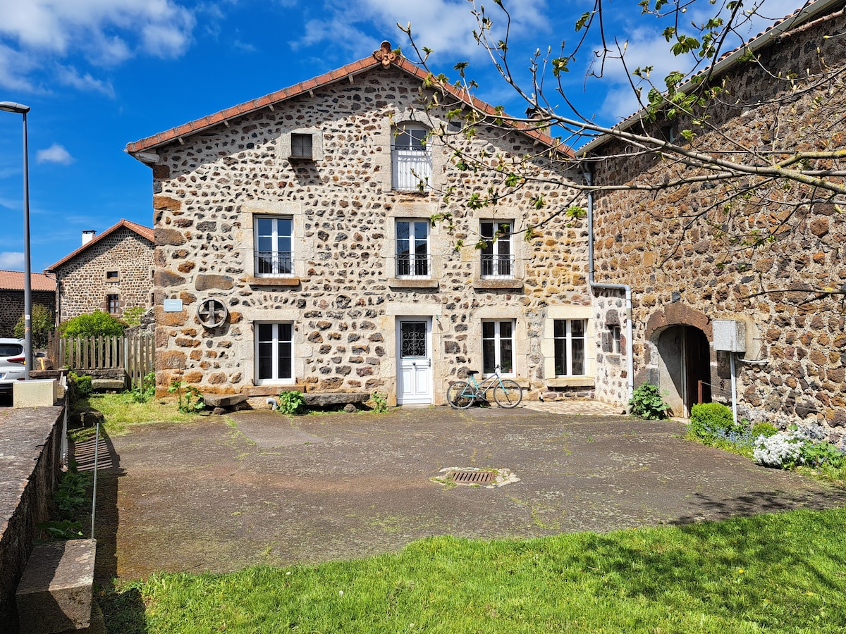 A stone facade building is presented with large windows offering natural light. A patio area is visible, featuring grass and paved sections. Bicycles are parked alongside the structure. The surroundings include additional stone buildings, enhancing the rural ambiance.