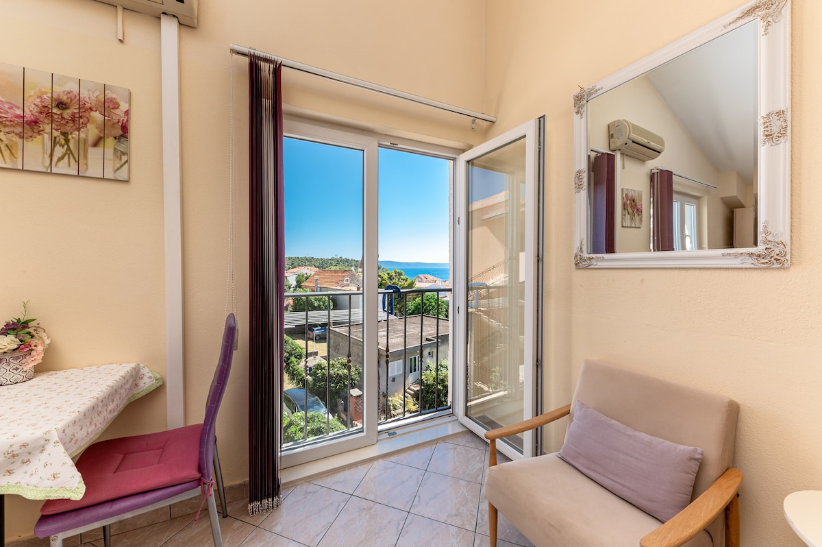 A compact dining area features a small table with a floral tablecloth and two chairs, one in deep purple and the other in natural wood. A large window offers a view of the sea and greenery outside, while natural light brightens the room.