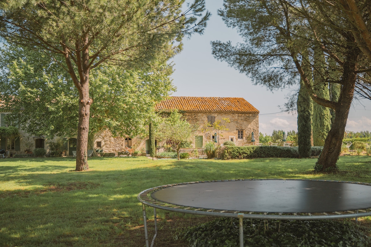 A spacious garden area is captured, featuring lush greenery and mature trees. In the foreground, a trampoline is visible on the grass. The stone house in the background is surrounded by greenery, showcasing a serene outdoor environment.
