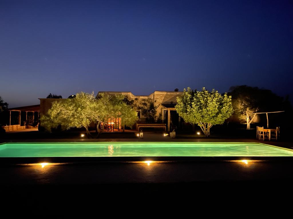 The villa is illuminated at night, showcasing a tranquil swimming pool that reflects the surrounding lights. Lush greenery and trees frame the pool area, creating a serene outdoor space. The structure of the villa is visible in the background, complementing the peaceful ambiance.