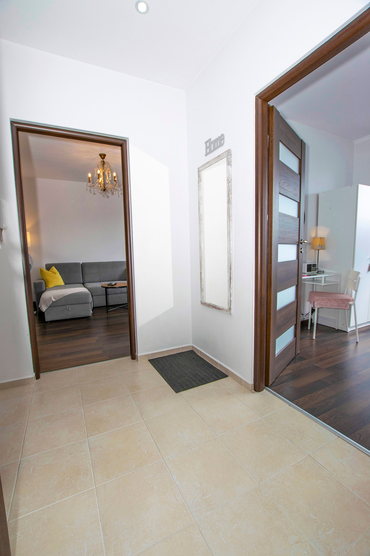 An inviting entryway is presented, featuring a light-colored tiled floor. The adjacent living space is visible, showcasing a gray sofa and a small coffee table. A decorative mirror and a door frame add to the welcoming ambiance, allowing natural light to brighten the area.