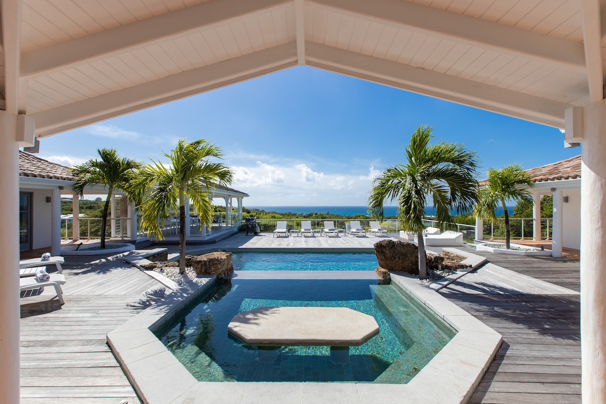 A dual pool area features a larger swimming pool and a smaller inset pool with a tiled table. Surrounded by a wooden deck, palm trees and native boulders enhance the tropical setting, creating a serene outdoor space with expansive views of the landscape.