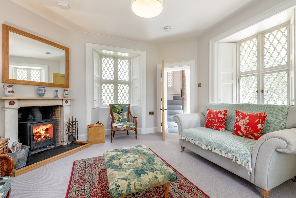 The inviting living area features a comfortable sofa with decorative cushions, arranged around a traditional fireplace with a warm fire. Light fills the space through diamond-patterned windows, highlighting the tasteful décor, a vintage armchair, and a decorative area rug.