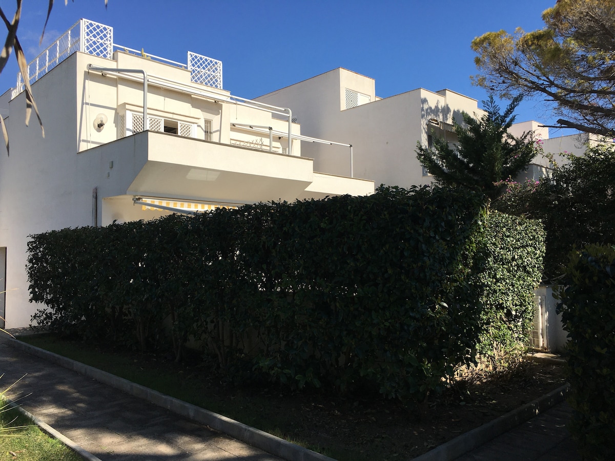 A modern white building is partially obscured by a tall, neatly trimmed hedge. The property features a balcony area above the entrance, with an outdoor railing visible. Clear blue skies provide a bright backdrop to the scene.