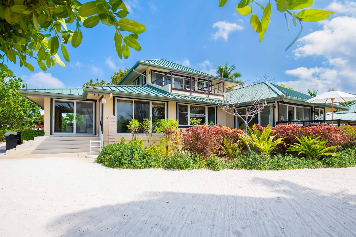 The villa features a modern design with a green metal roof and large windows, surrounded by vibrant tropical landscaping. The sandy beach leads directly to the entrance, while outdoor seating areas and palm trees create a refreshing coastal atmosphere.