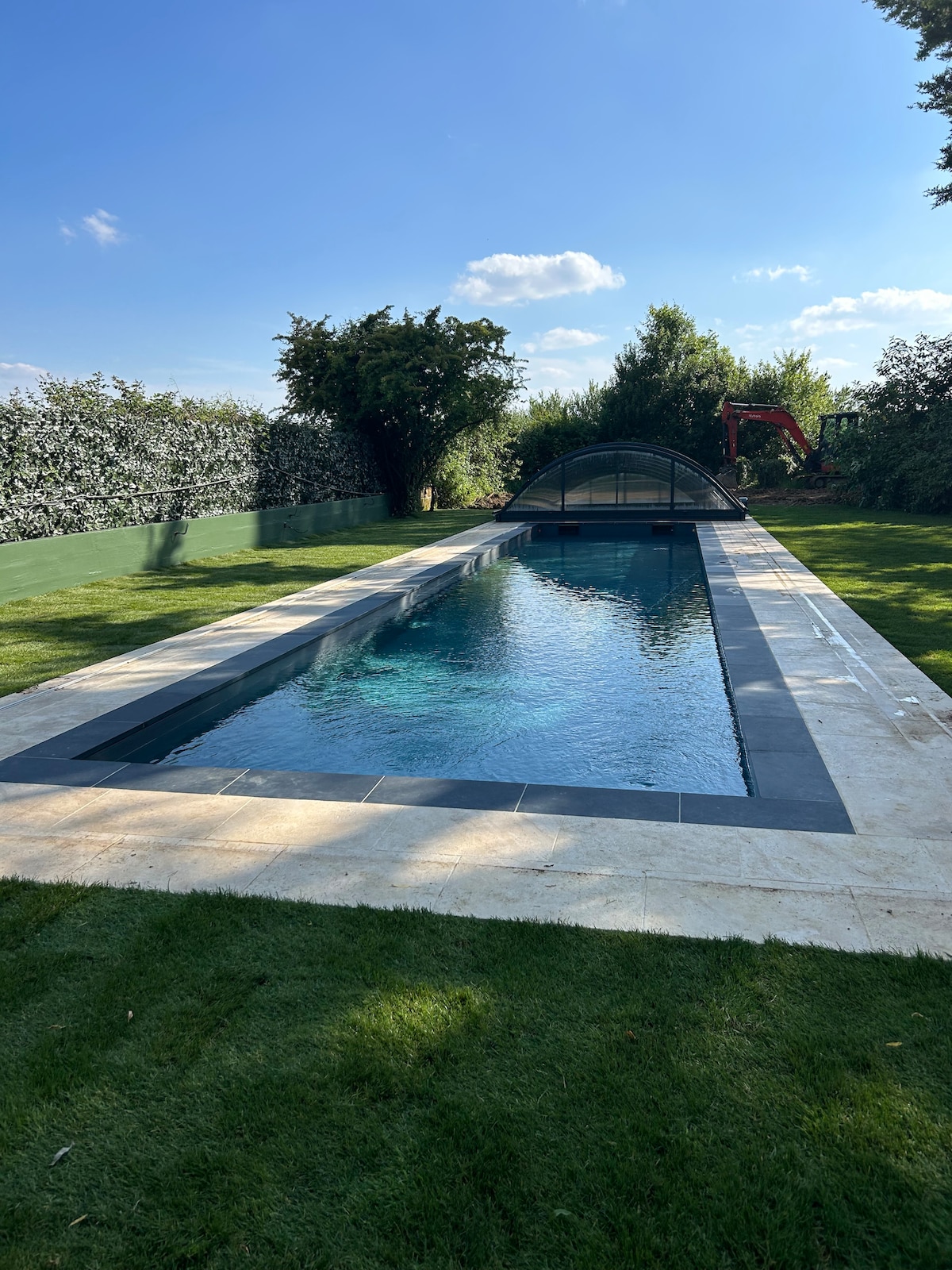 A swimming pool measuring 15 meters by 3 meters is surrounded by a well-maintained grass lawn. The pool features a clear, inviting water surface reflecting the sky. A safety cover is positioned over the pool, and lush greenery provides privacy along the perimeter.