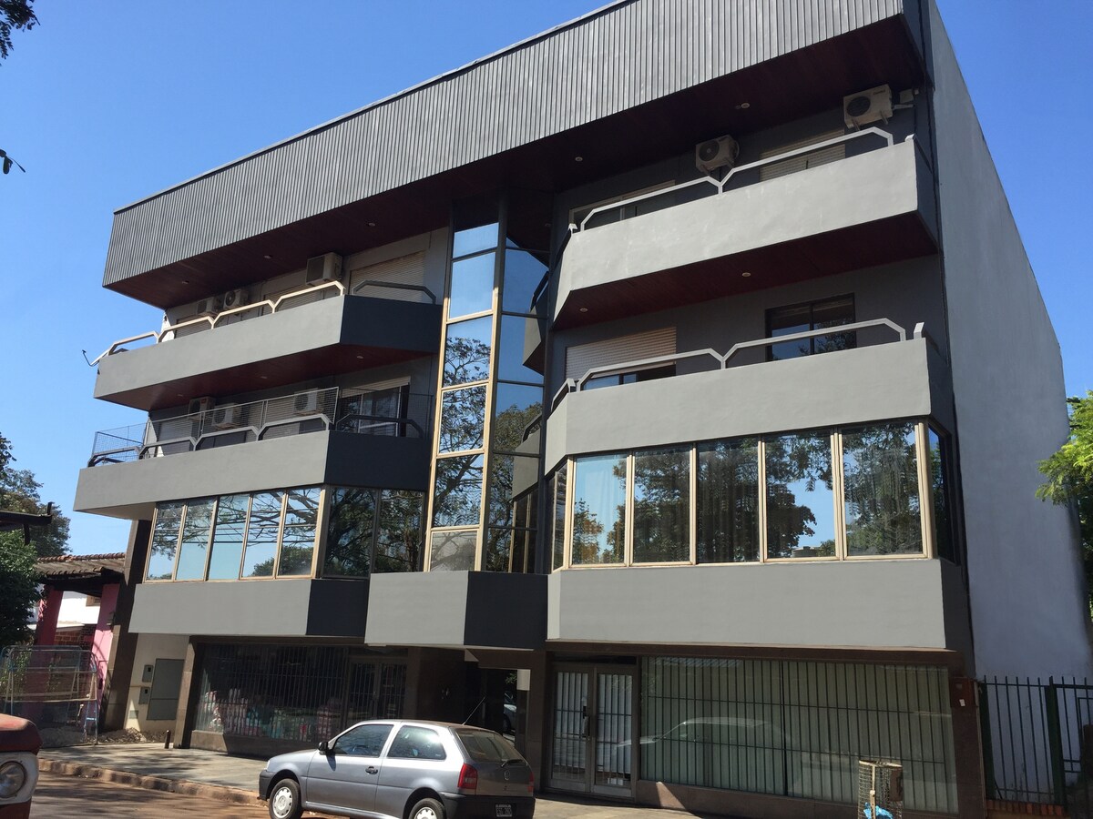 A modern building features a minimalist design with large glass windows on the upper levels. Several air conditioning units are visible on the exterior walls. Nearby, an off-street parking area accommodates vehicles, and trees provide greenery around the structure.