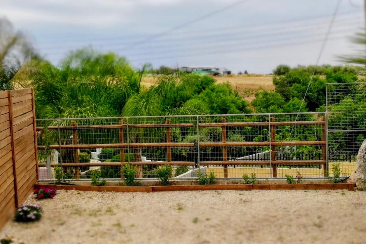 A private garden area is visible, featuring gravel grounds bordered by wooden fencing. Lush greenery, including palm trees and shrubs, can be seen beyond the fence, providing a serene backdrop. The scene is complemented by a well-maintained landscape with flowers at the garden edge.
