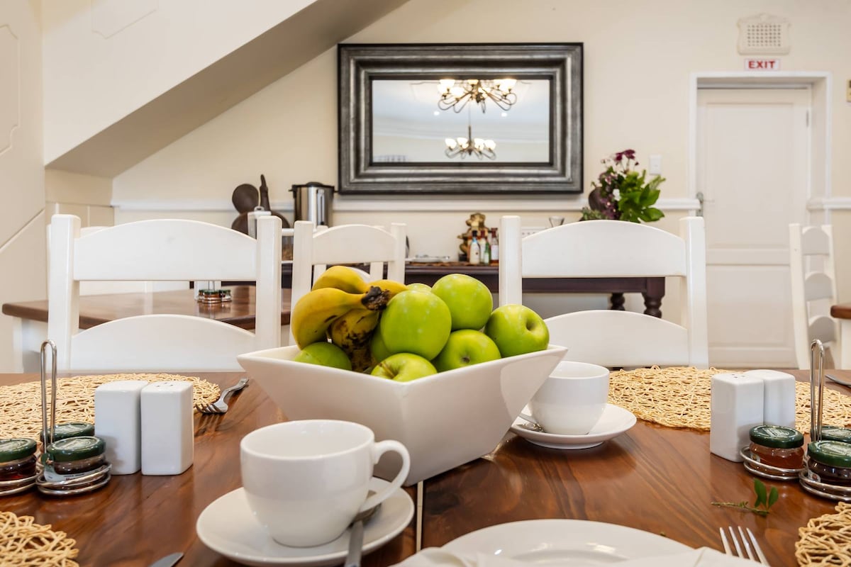 A dining table is set with white cups and saucers, accompanied by jars of jam. A centerpiece bowl filled with fresh bananas and green apples adds a touch of natural color. A large decorative mirror is mounted on the wall, reflecting the inviting space.