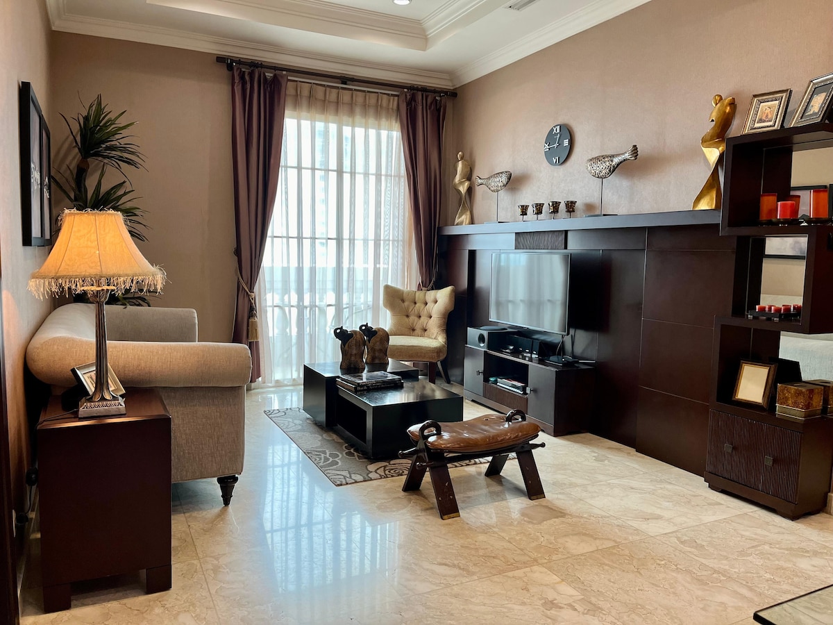 The living room is designed with a neutral colour palette, featuring a comfortable sofa and an elegant armchair. A coffee table sits in the center, surrounded by decorative accents. Natural light enters through large glass doors, which lead to a balcony, enhancing the inviting space.