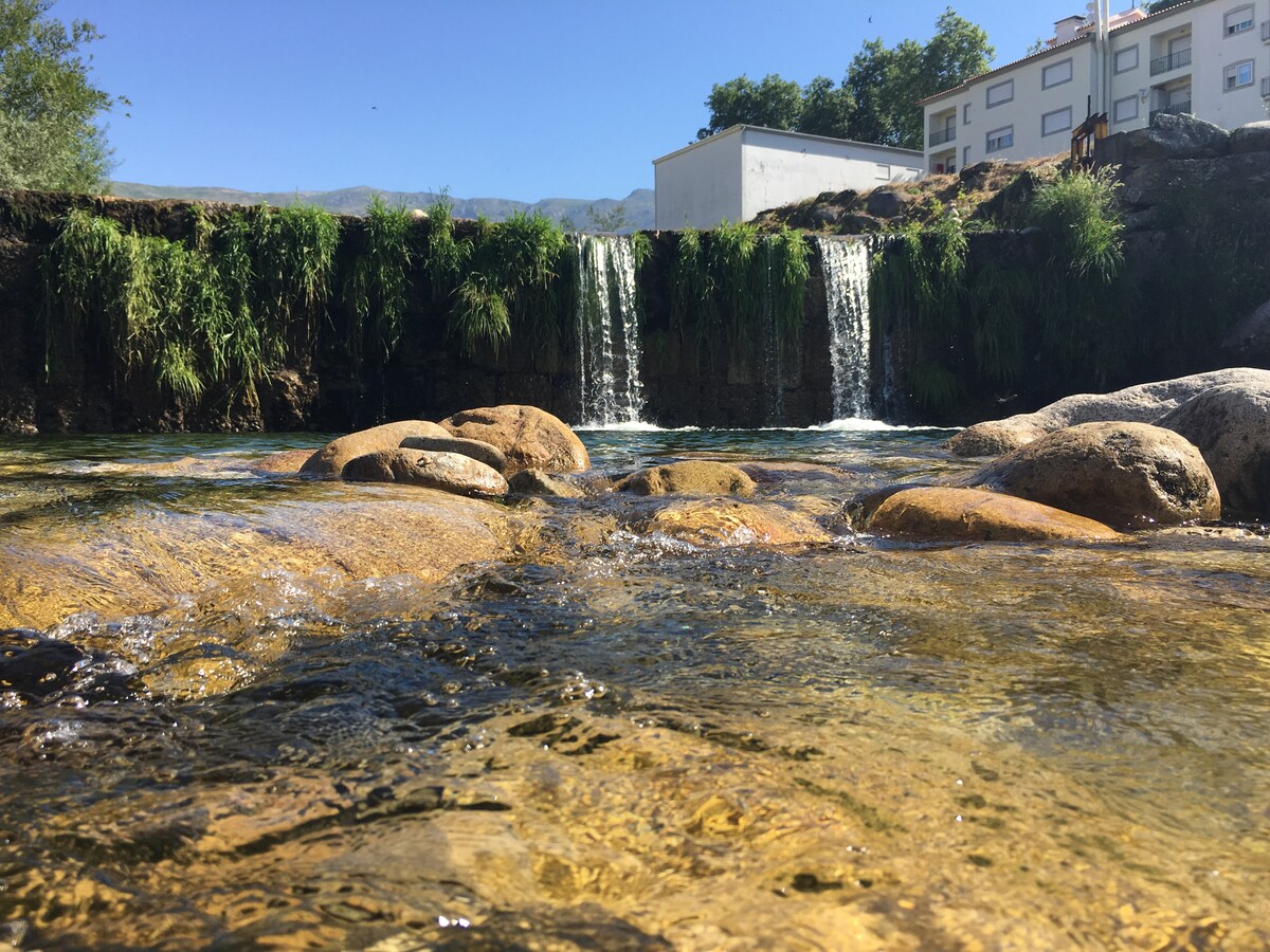A serene natural setting is depicted, featuring clear water flowing over rocks and creating a gentle waterfall. Lush green grasses line the water's edge, while a building can be seen in the background, showcasing a harmonious blend of nature and structure.
