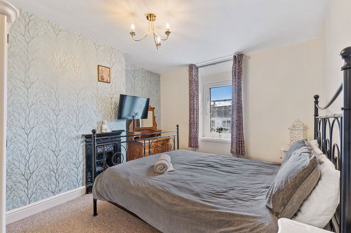 A spacious bedroom features a large dark bed frame with neutral linens. A television is mounted on the wall across from the bed, and a wooden dresser is positioned beside the window. Light enters through the window, partially covered by a patterned curtain.