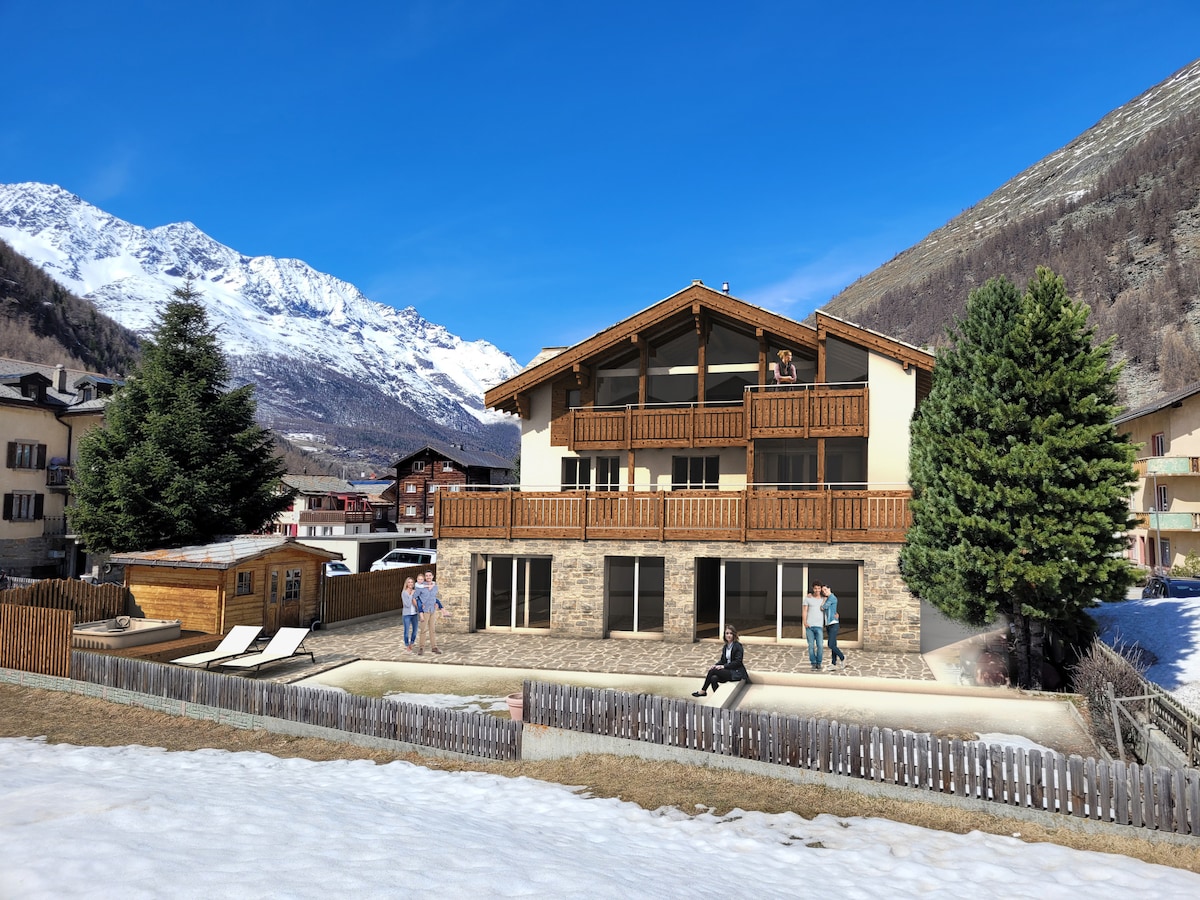The exterior of a spacious chalet is presented, featuring a dual-level structure with wooden balconies that overlook scenic mountainous views. A patio area with lounge chairs is visible, surrounded by landscaped grounds. Snow-covered areas and nearby trees complete the tranquil setting.