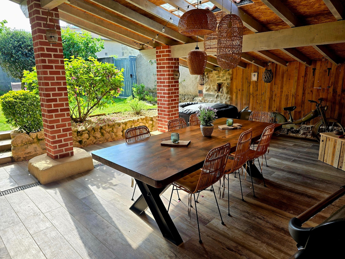An outdoor dining area features a large wooden table accompanied by several woven chairs. Natural light filters through the beams above. Lush greenery surrounds the space, adding a touch of nature. A bicycle is visible in the background, enhancing the relaxed atmosphere.