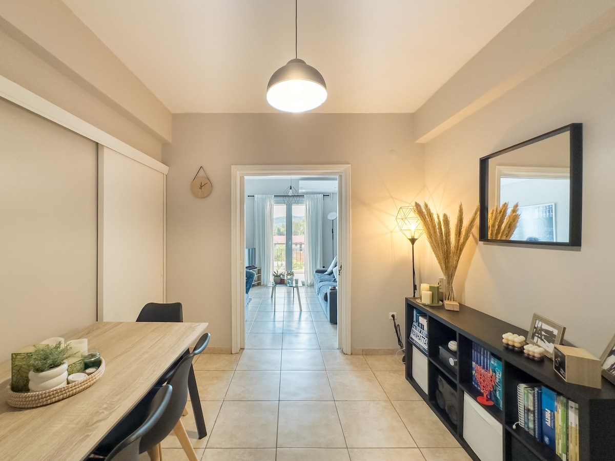 The interior space features a dining area with a wooden table and black chairs. A decorative shelf is visible, displaying various plants and books. Light enters through an open doorway leading to a living area, with soft lighting fixtures enhancing the overall ambiance.