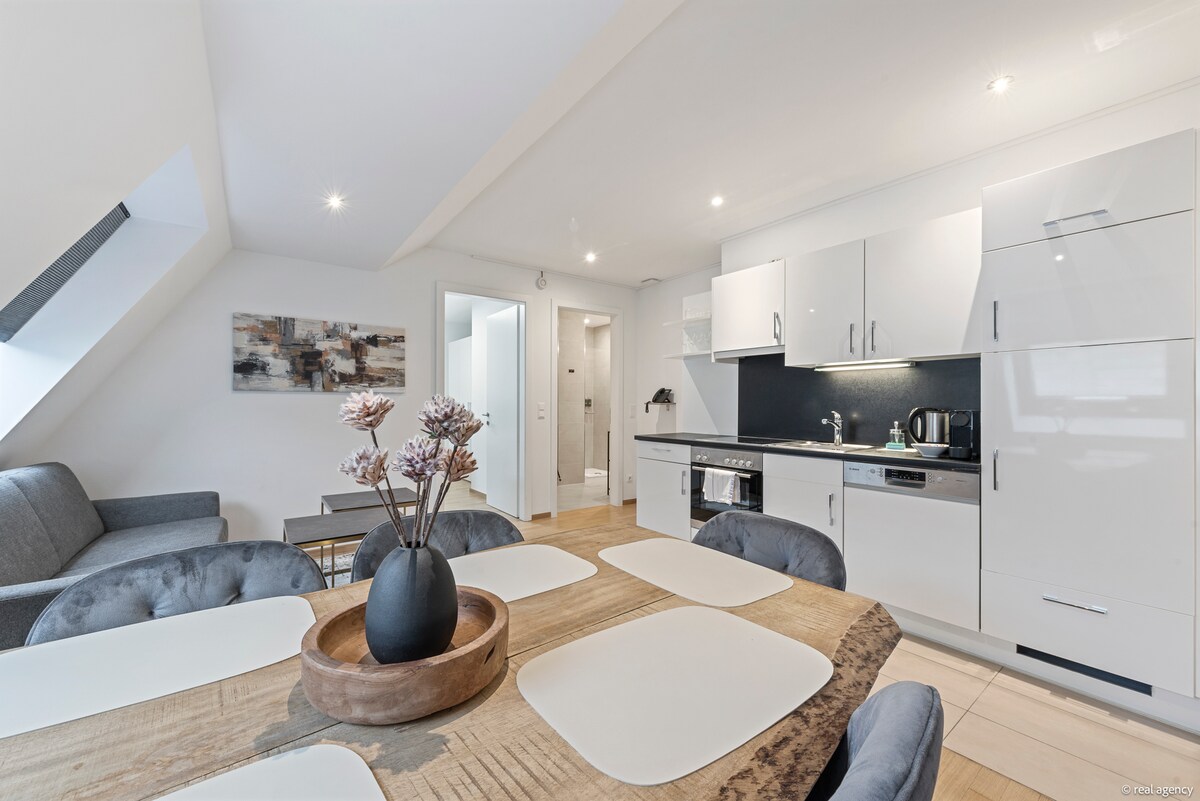 An open kitchen is featured, equipped with modern appliances, including an oven and coffee maker. A dining table accommodates eight, surrounded by plush chairs. A cozy couch is positioned nearby, and decorative elements include a coffee table with a wooden bowl and a floral arrangement.