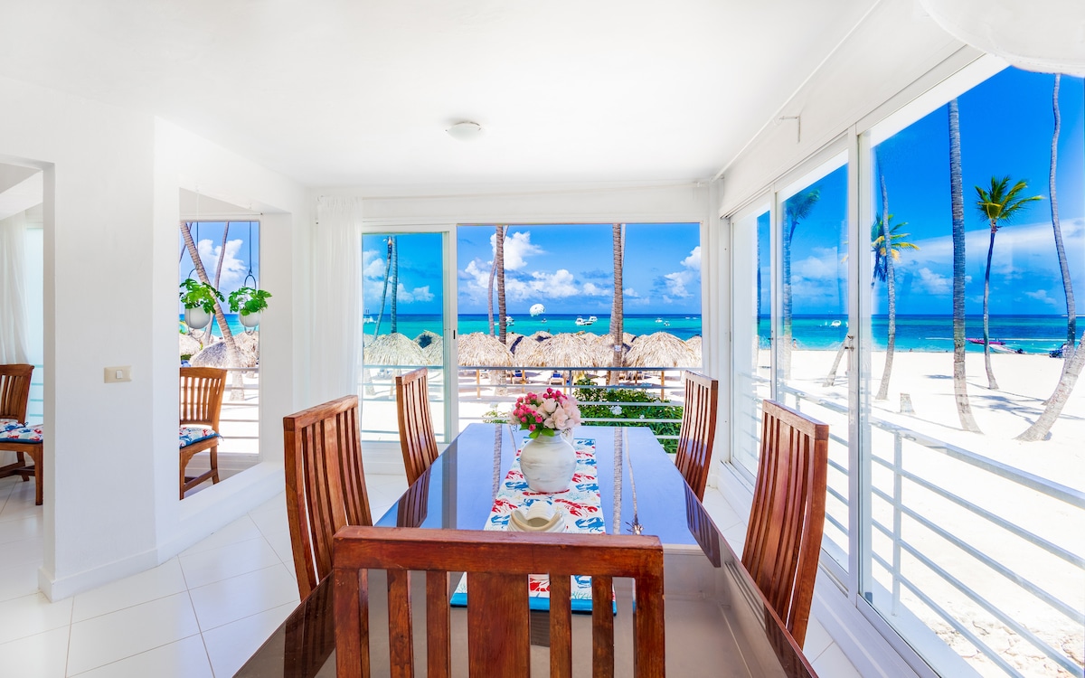A bright dining area is seen with a large wooden table surrounded by matching chairs. Floor-to-ceiling windows offer unobstructed views of the beach and ocean, with palm trees swaying gently in the background. Natural light fills the space, creating a vibrant atmosphere.