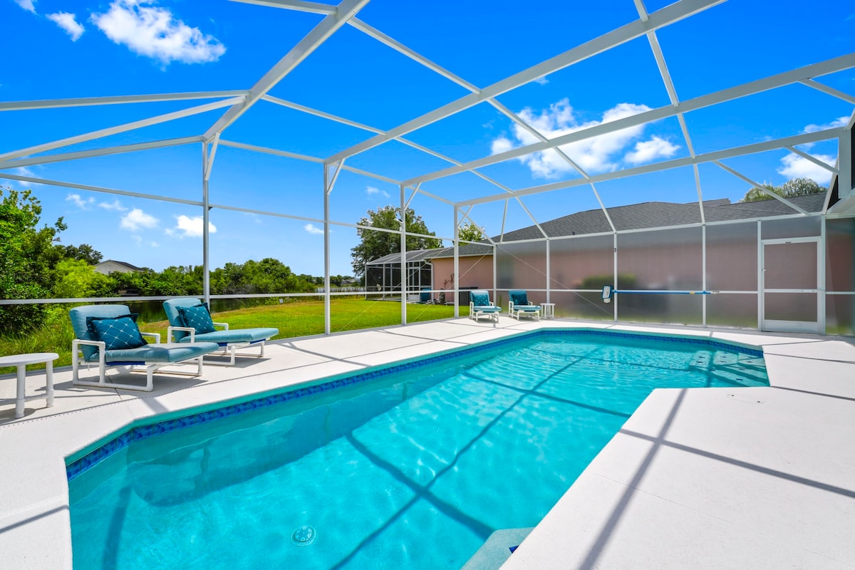 A spacious pool area is enclosed by a screened lanai, featuring clear blue water and a fresh, sunlit environment. Several lounge chairs are arranged thoughtfully around the pool, inviting relaxation. The lush greenery of the surrounding landscape is visible beyond the enclosure.