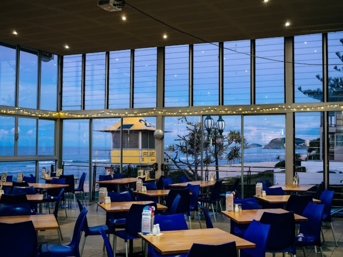 A spacious dining area is furnished with wooden tables and blue chairs, offering a comfortable environment for meals. Large windows provide views of the ocean and a nearby lifeguard station, enhanced by the soft evening light. String lights add a warm ambiance to the setting.