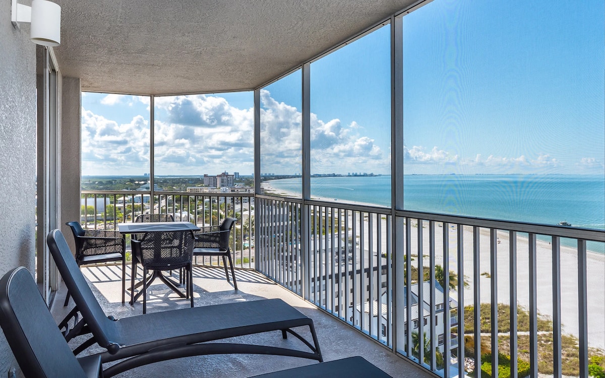 A spacious screened balcony features a dining table with four chairs and two lounge chairs, offering panoramic views of the Gulf of Mexico. The area is flooded with natural light, enhancing the serene atmosphere and providing a perfect spot for relaxation.