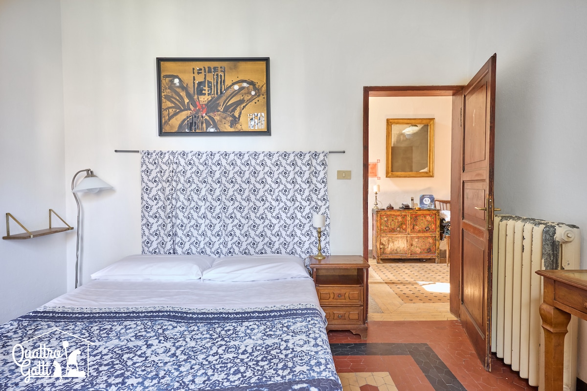A bright guest room features a spacious bed with a decorative blue and white patterned coverlet. A framed artwork hangs above the headboard, while wooden furniture adds warmth. A door leads to another area, enhancing the open feel of the space.