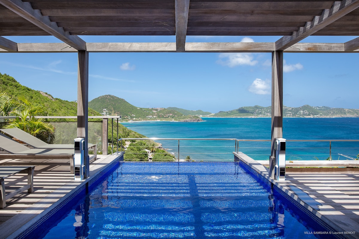 An infinity pool is positioned at the forefront, showcasing clear blue water blending into the ocean. Sun loungers are arranged along the deck, while the view of Lorient Bay and the lush hillsides creates a serene backdrop under a bright sky.