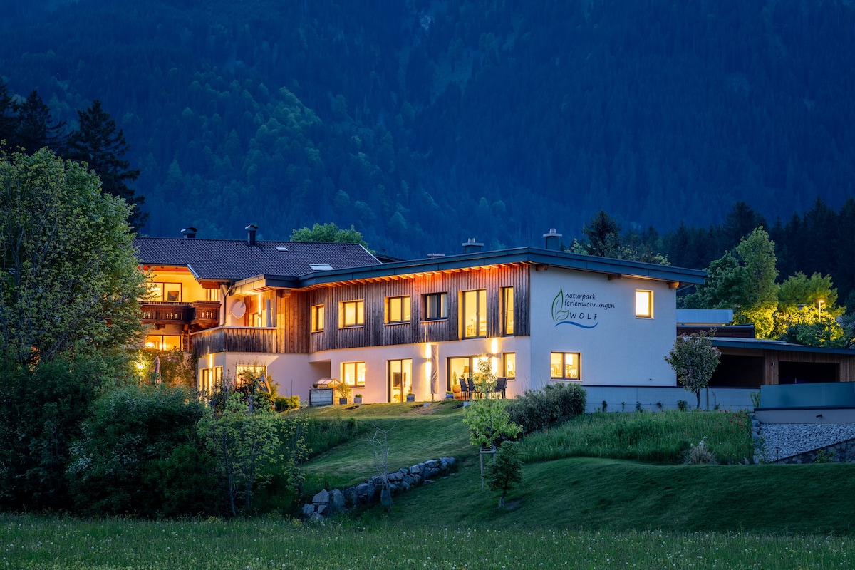 A modern multi-story building is set against a backdrop of mountains, illuminated warmly at night. Windows provide glimpses of light from within. Lush greenery surrounds the structure, enhancing the inviting scene. A well-maintained lawn leads to the entrance, contributing to a welcoming atmosphere.