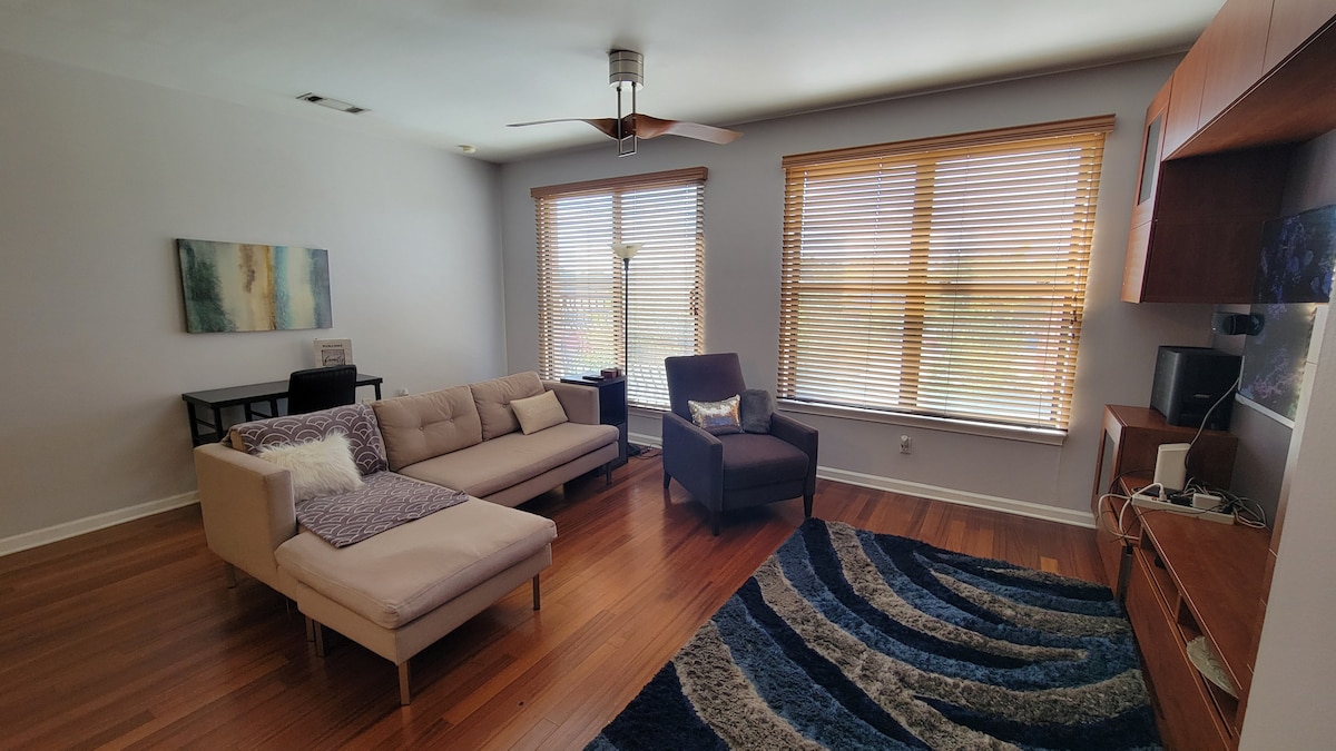 A spacious living area features hardwood floors and large windows with blinds, allowing natural light to fill the space. A comfortable sectional sofa is paired with an armchair, creating a cozy seating arrangement. A decorative rug with wavy patterns adds visual interest.