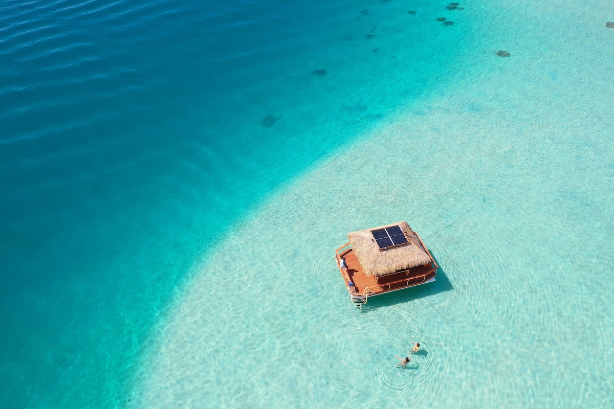 A floating structure with a thatched roof and solar panels is seen in the clear, calm waters. Two individuals are swimming nearby, with light rays visible beneath the turquoise surface, showcasing the serene aquatic environment.