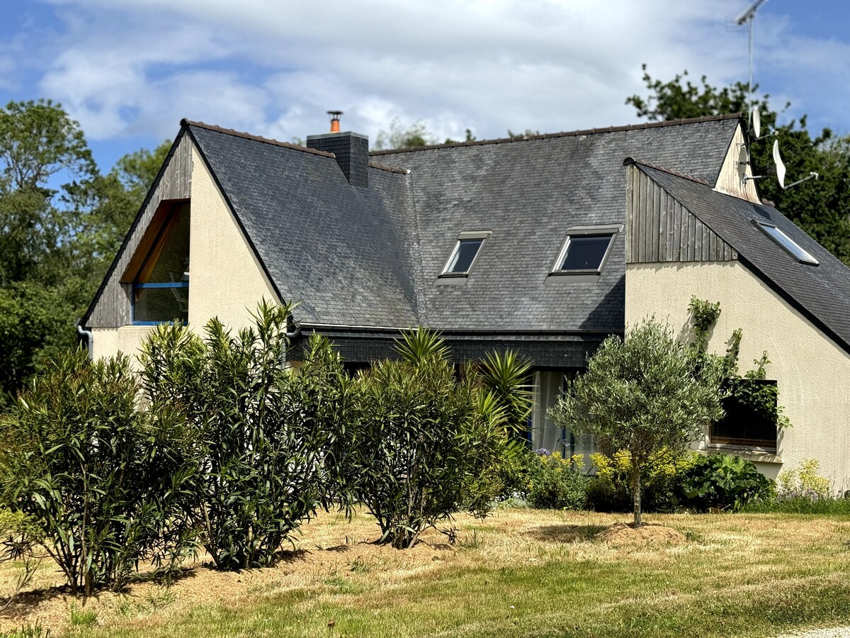 A two-story house features a sloping roof with multiple skylights. Surrounding greenery includes shrubs and trees, providing a natural frame. The exterior is painted in light tones, with elements of wood accents visible. A peaceful garden area is presented in the foreground.