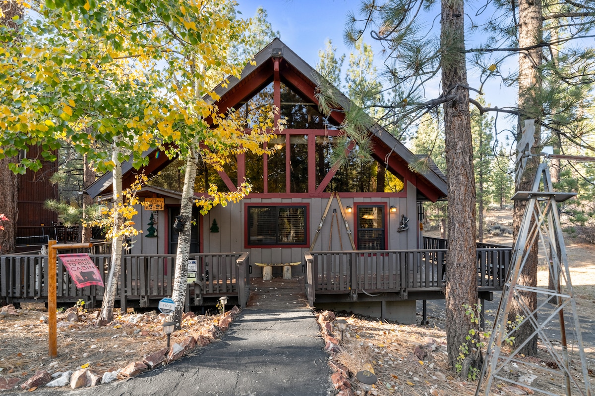 The cabin’s exterior features a welcoming facade with large windows and a peaked roof. Surrounded by tall trees, the entrance is approached via a paved pathway. A wooden deck with railings is visible, inviting guests to relax in a natural setting.