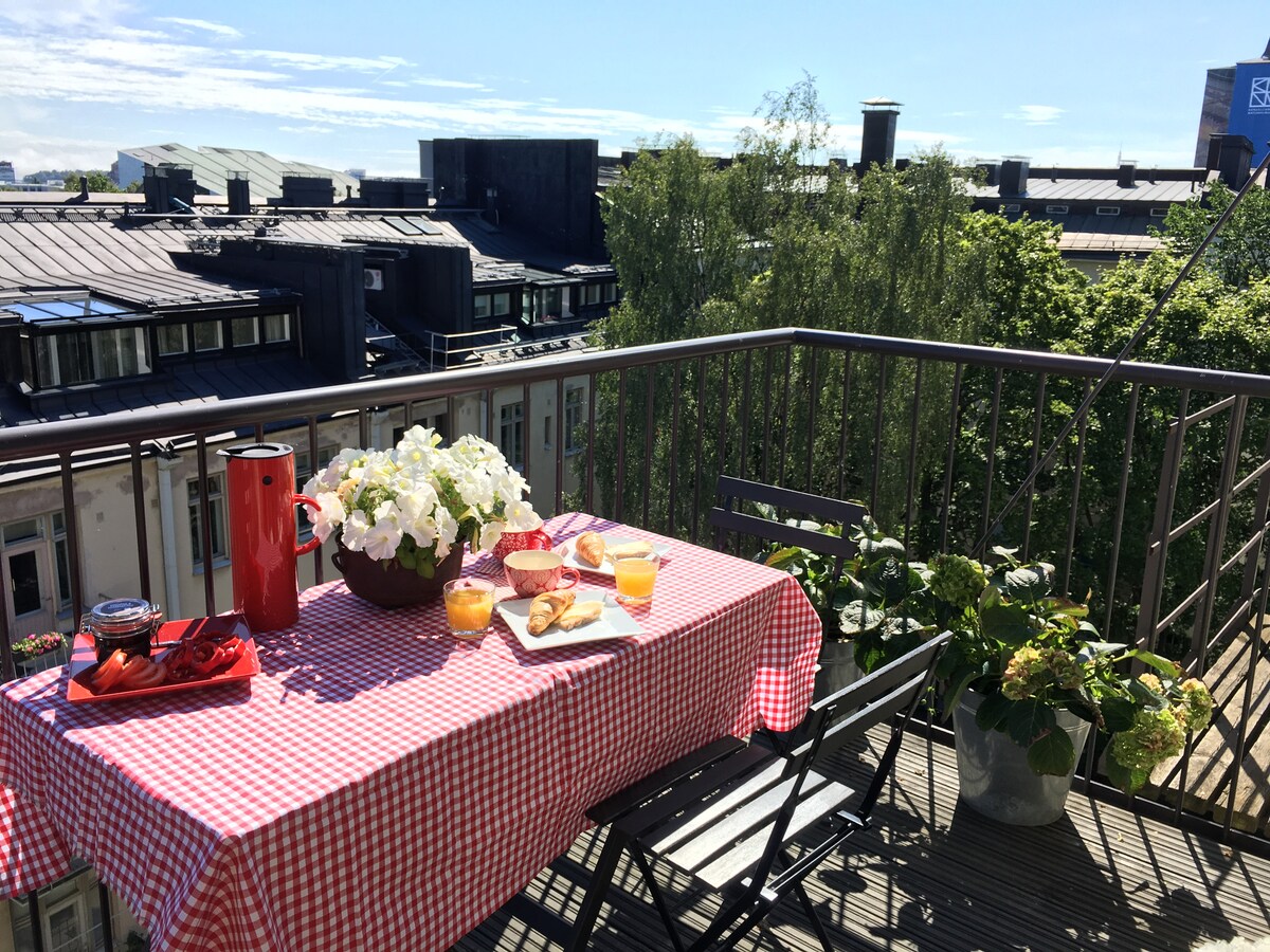 A table covered with a red and white checkered tablecloth is set for breakfast on the east-facing balcony. Fresh flowers and breakfast items are arranged, accompanied by drinks. Surrounding greenery and rooftops create a peaceful view, inviting relaxation in the outdoor space.
