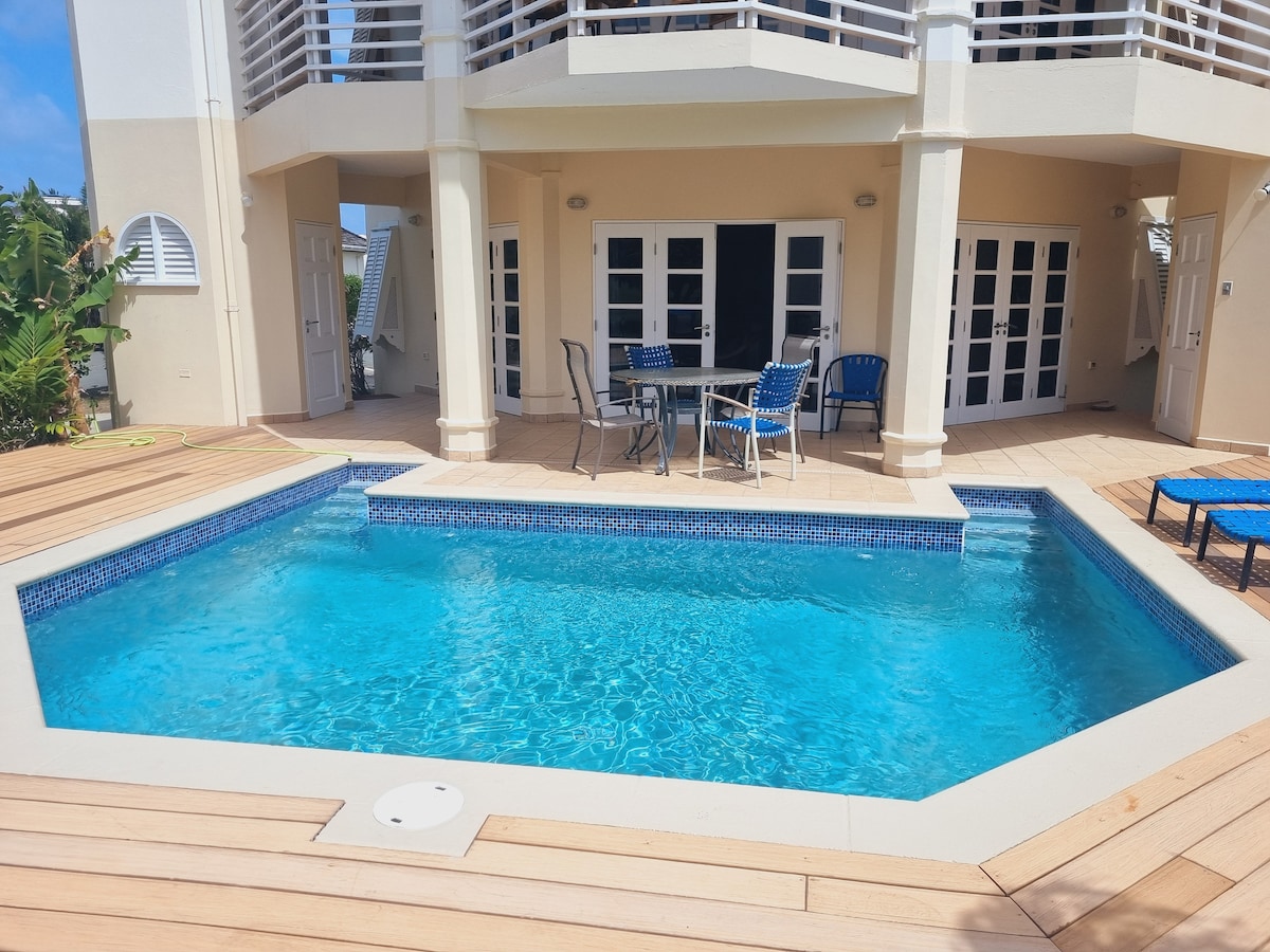 A clear plunge pool is situated within a wooden deck area, surrounded by lounge chairs. The adjacent outdoor dining set includes four metal chairs with a round table. Multiple large glass doors offer access to the villa's interior, allowing for natural light.