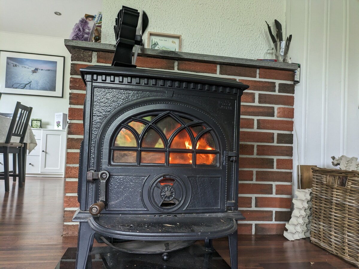 A black cast iron fireplace is positioned against a brick wall, with flames visibly flickering inside. The fireplace features an arched window design and a sturdy base. The surrounding area is decorated with a wicker basket and kitchen utensils displayed on a shelf above.