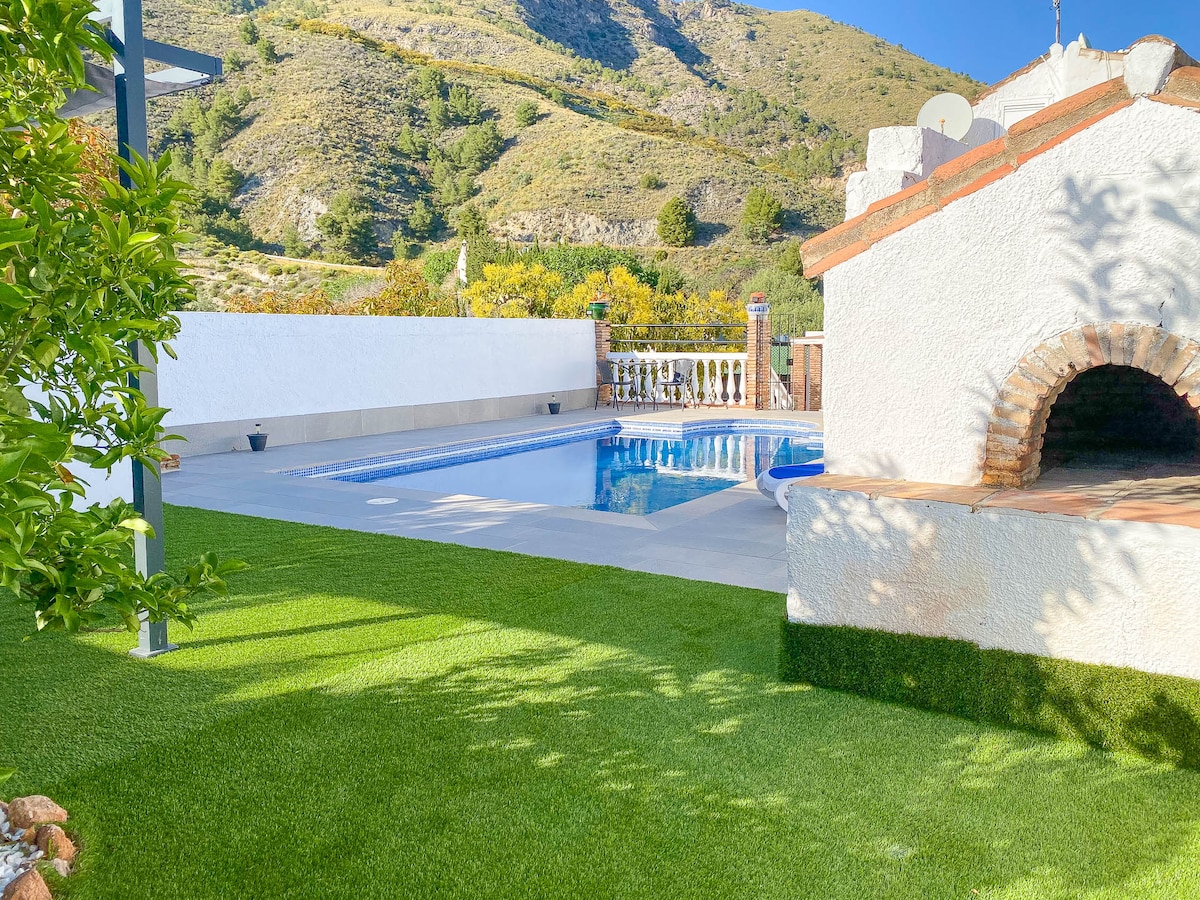 A private pool is set among landscaped grounds, with well-maintained artificial grass surrounding it. A stone barbecue area is visible near the pool, and the serene backdrop features rolling hills, demonstrating a natural and tranquil environment.